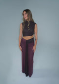 A woman modeling a sleeveless, backless dark brown crop top and a matching deep plum purple maxi skirt, standing against a solid light gray studio background.