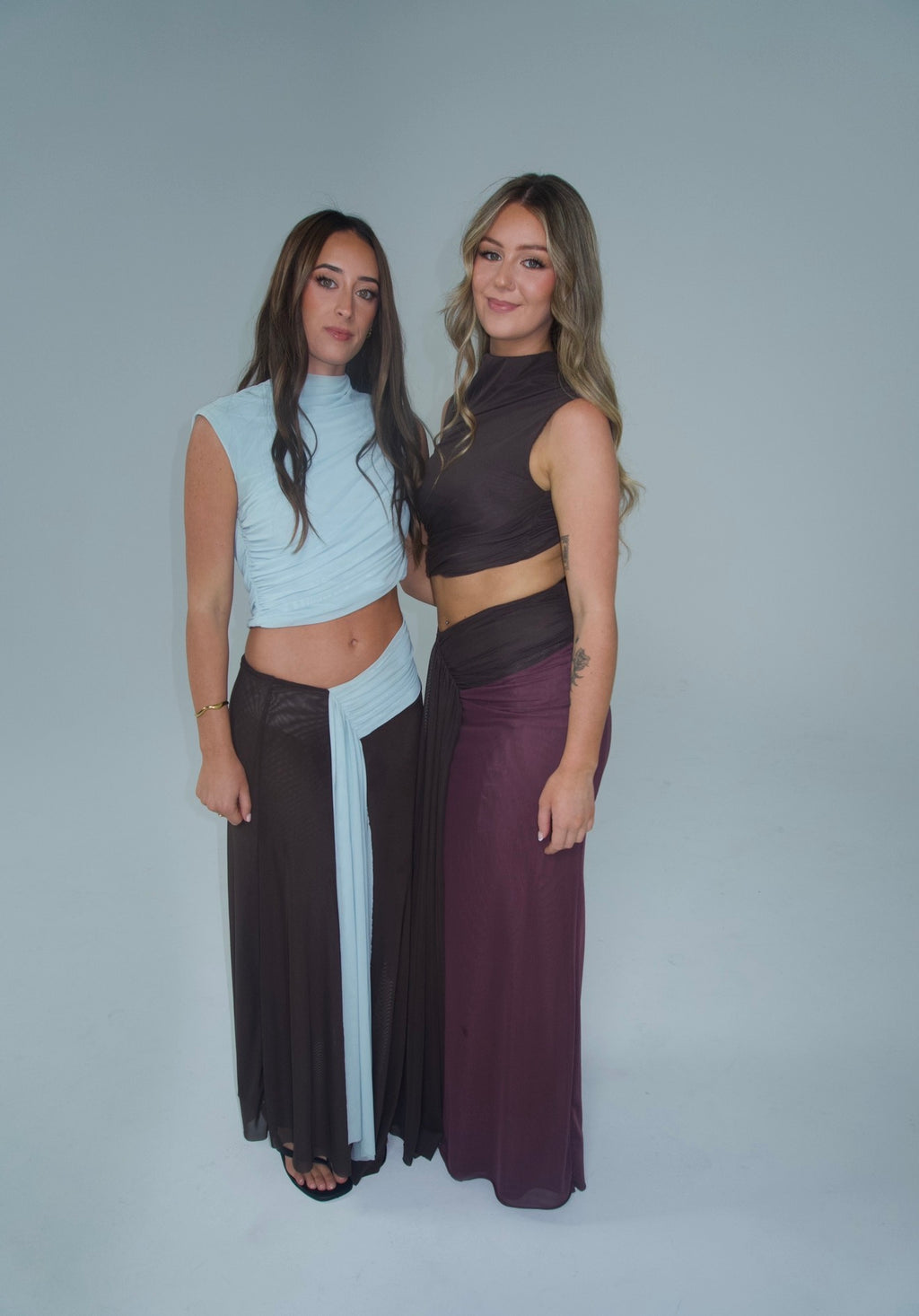 Two women modelling coordinated mesh two-piece outfits, one in light blue and chocolate brown and the other in chocolate brown and plum, standing together against a light grey studio background.