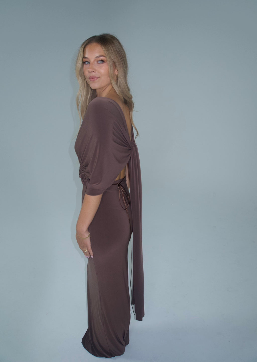 A woman modeling a floor-length, dark brown off-the-shoulder gown with a deep V-neckline and gathered waist details, set against a solid light gray studio background.