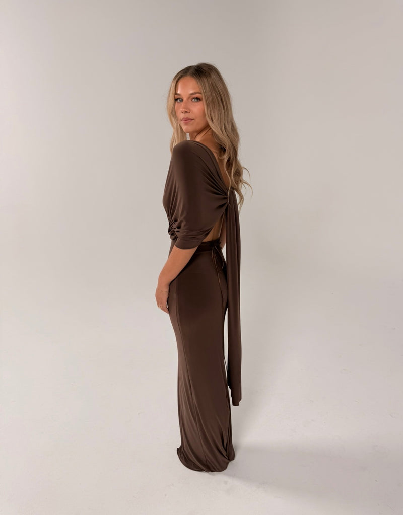 A woman modeling a floor-length, dark brown off-the-shoulder gown with a deep V-neckline and gathered waist details, set against a solid light gray studio background. - @indiabluegranger
