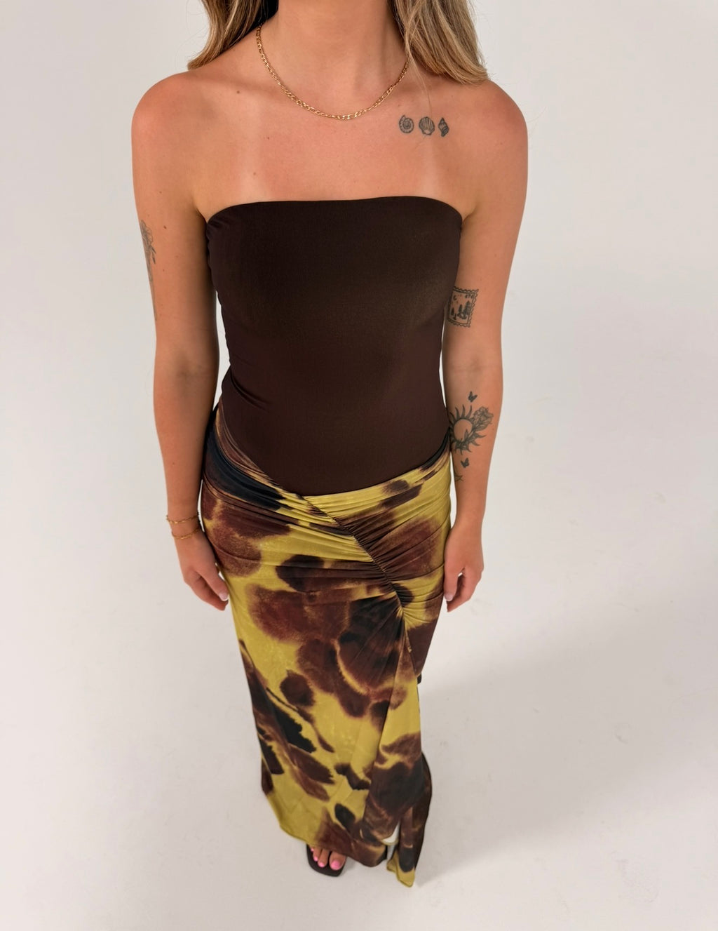 Woman wearing a strapless chocolate brown fitted top with a draped maxi skirt featuring an abstract olive green, brown and black print, photographed against a light grey studio background.