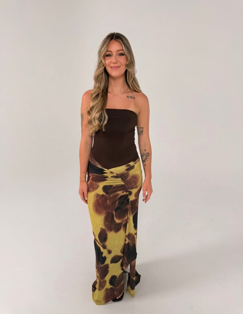 Woman wearing a strapless chocolate brown fitted top with a draped maxi skirt featuring an abstract olive green, brown and black print, photographed against a light grey studio background.