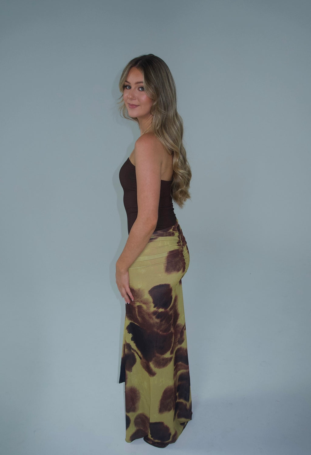 Alt text: Woman wearing a strapless chocolate brown fitted top with a draped maxi skirt featuring an abstract olive green, brown and black print, photographed against a light grey studio background.