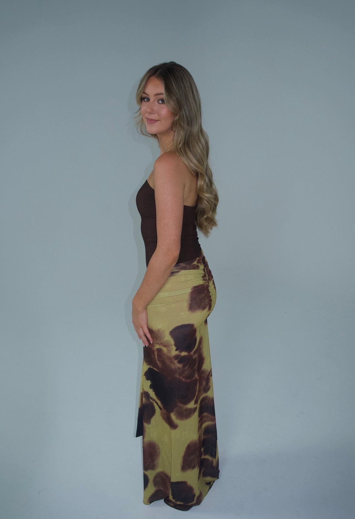 Alt text: Woman wearing a strapless chocolate brown fitted top with a draped maxi skirt featuring an abstract olive green, brown and black print, photographed against a light grey studio background.