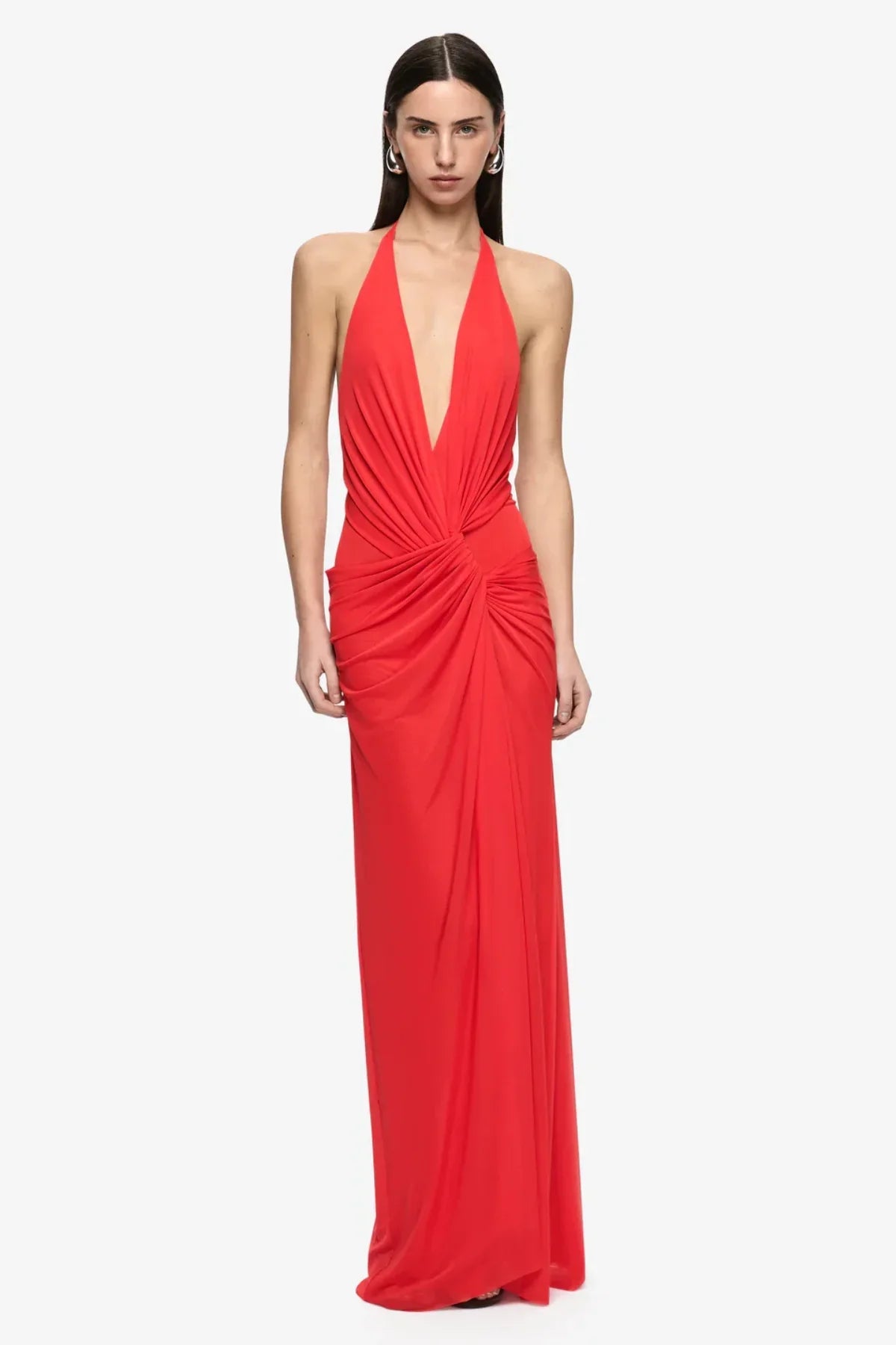 Woman wearing the Bec + Bridge Xanthi V Neck Maxi Dress in Flame, featuring a plunging halter neckline, gathered waist detailing, flowing draped skirt and dramatic thigh-high split, styled with gold heels against a neutral background.
