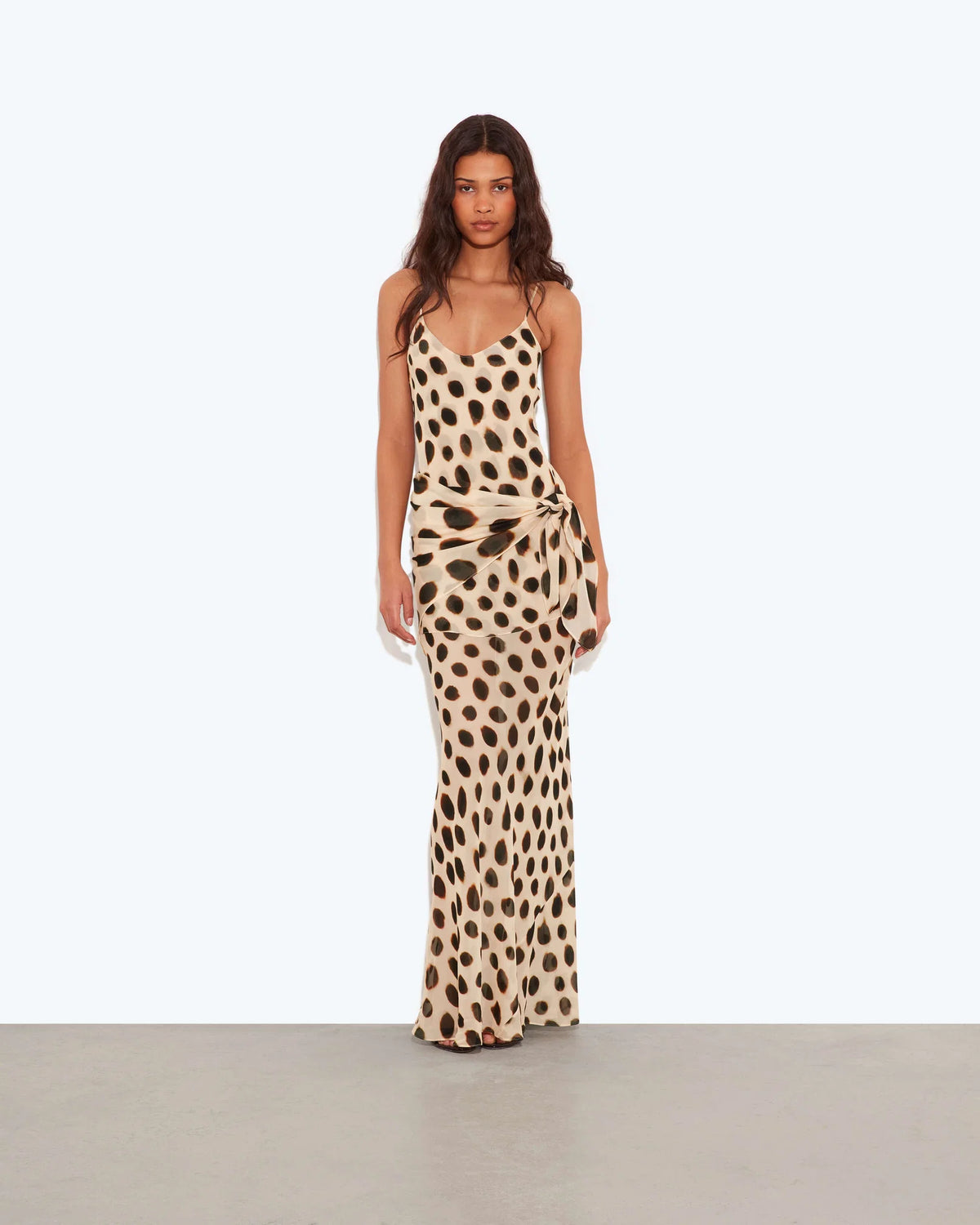 A woman modeling a sleeveless maxi dress with a beige and brown animal print pattern, standing against a plain light gray studio background.