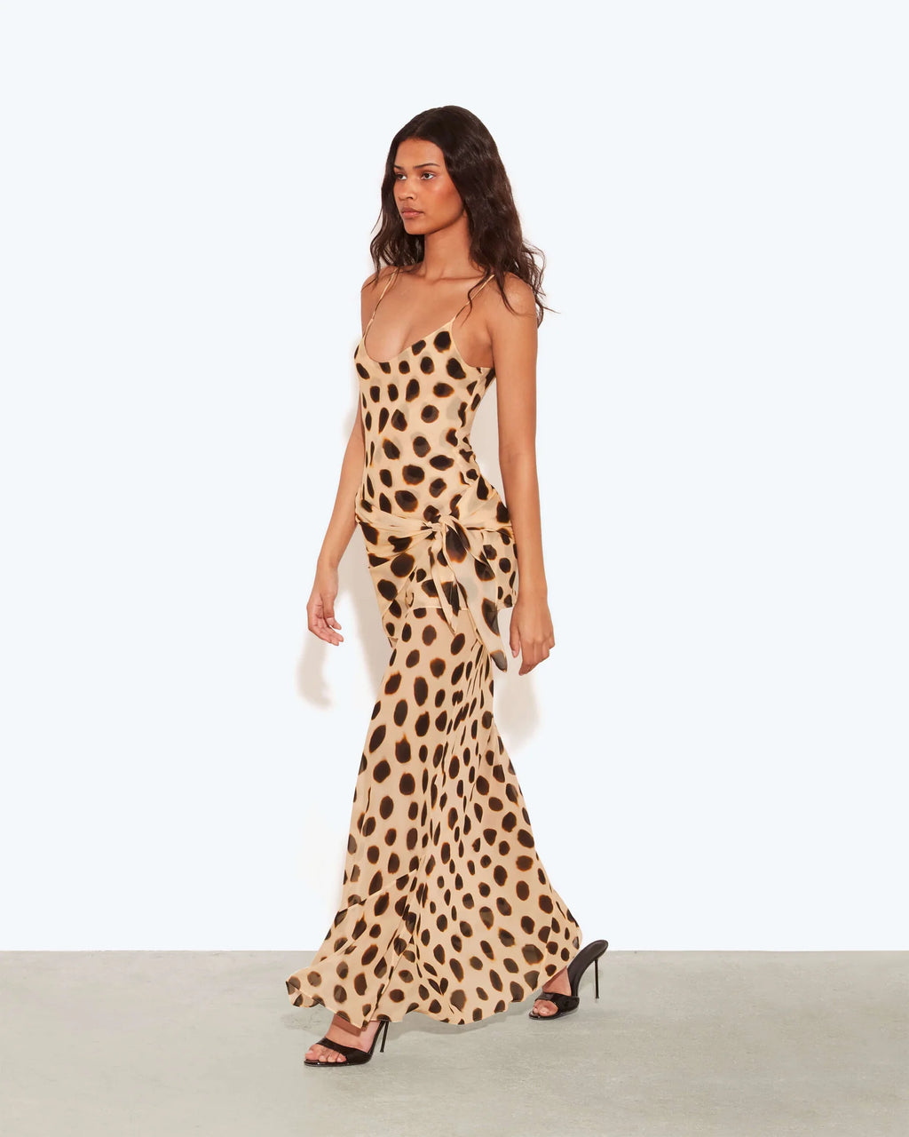 A woman modeling a sleeveless maxi dress with a beige and brown animal print pattern, standing against a plain light gray studio background.