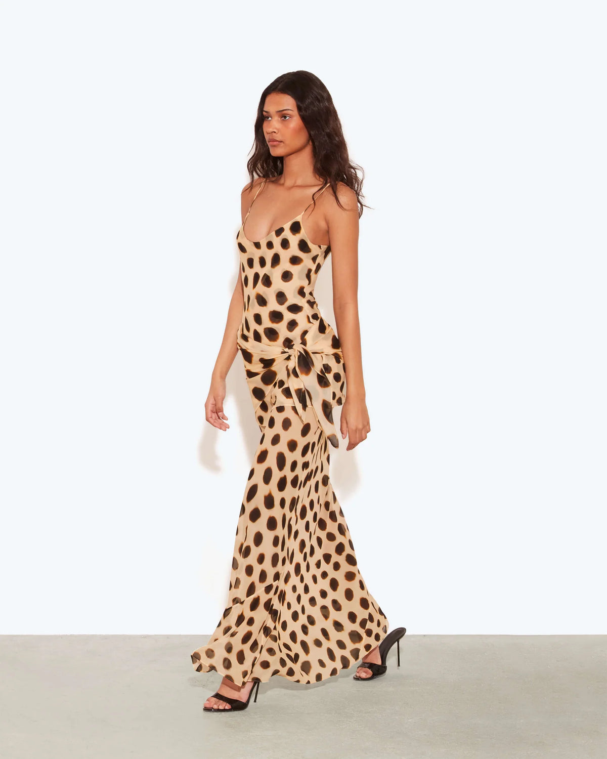 A woman modeling a sleeveless maxi dress with a beige and brown animal print pattern, standing against a plain light gray studio background.