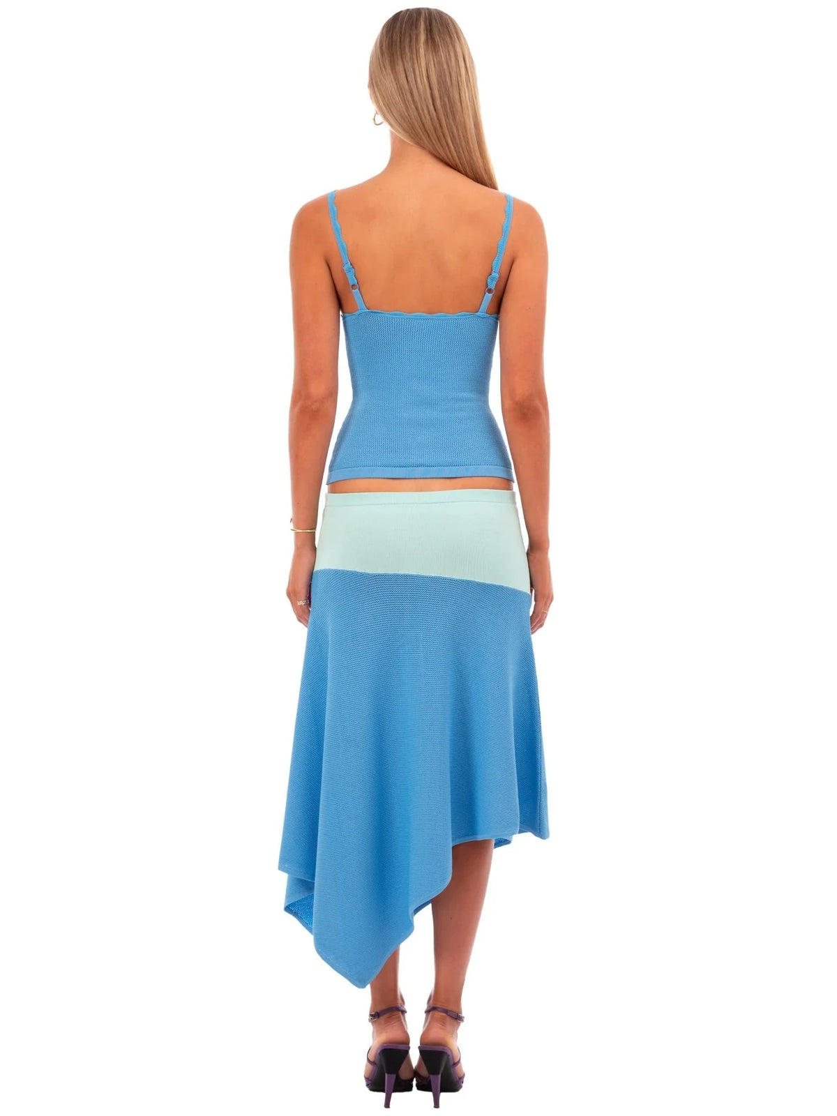 A woman shown from the back wearing a color-blocked knit two-piece set with a light blue crop top and an asymmetrical midi skirt in light blue and pale mint green, paired with black high heels on a white background.