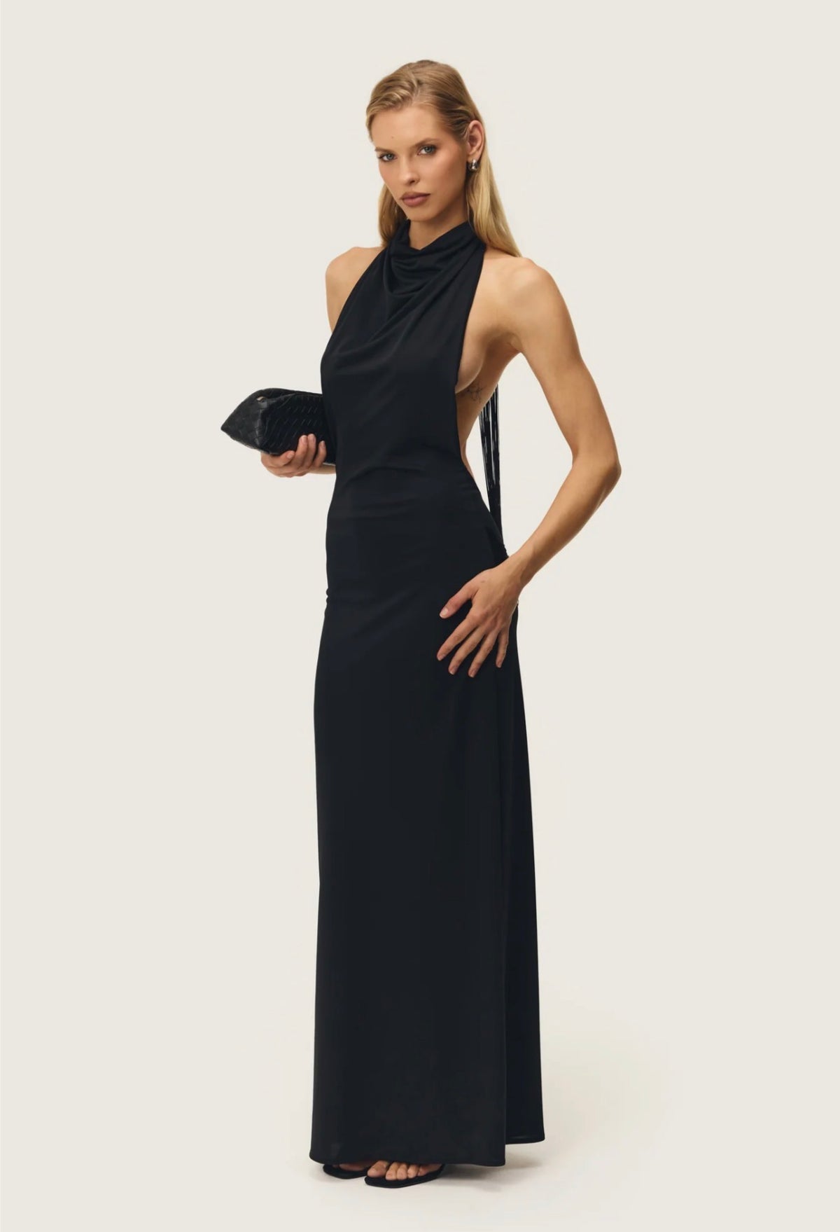 Model wearing the Arcina Ori Jean Dress in black, featuring a draped high halter neckline, open sides, flowing back tassels and a sleek floor-length silhouette.