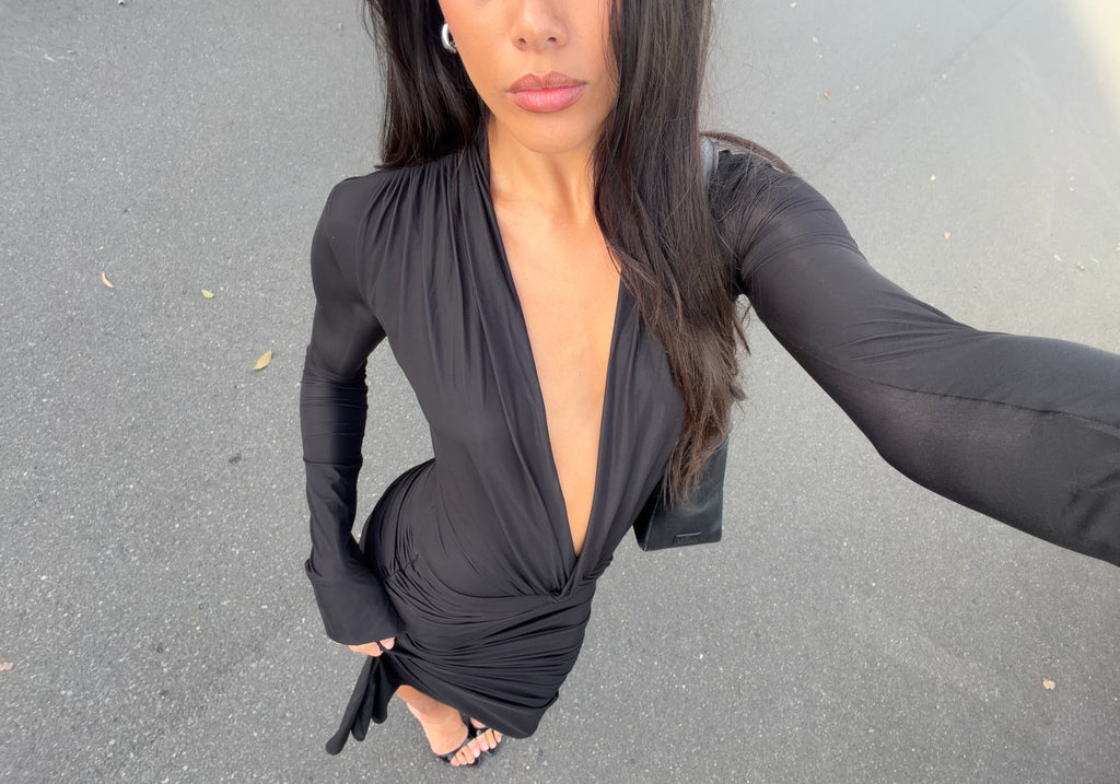 Woman wearing a black long-sleeve dress with a plunging neckline on a gray pavement background.