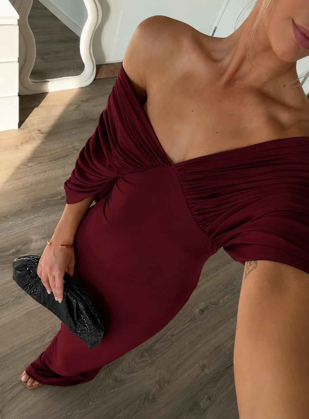 Woman in a burgundy off-shoulder dress holding a black clutch on a wooden floor.