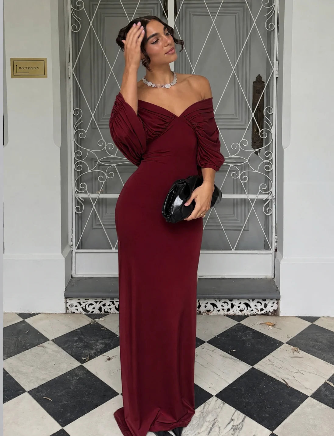 A woman wearing a floor-length off-the-shoulder burgundy maxi dress with ruched sleeves, holding a black clutch bag, standing in front of a white door with a metal security grille on a black and white checkered floor.