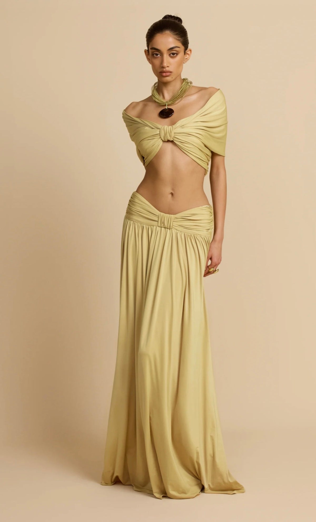 Model wearing the Alessia Set in a soft golden hue, featuring an off-shoulder ruched crop top with a gathered centre and a matching low-waisted, floor-length maxi skirt with flowing draped fabric, styled against a neutral beige studio background.