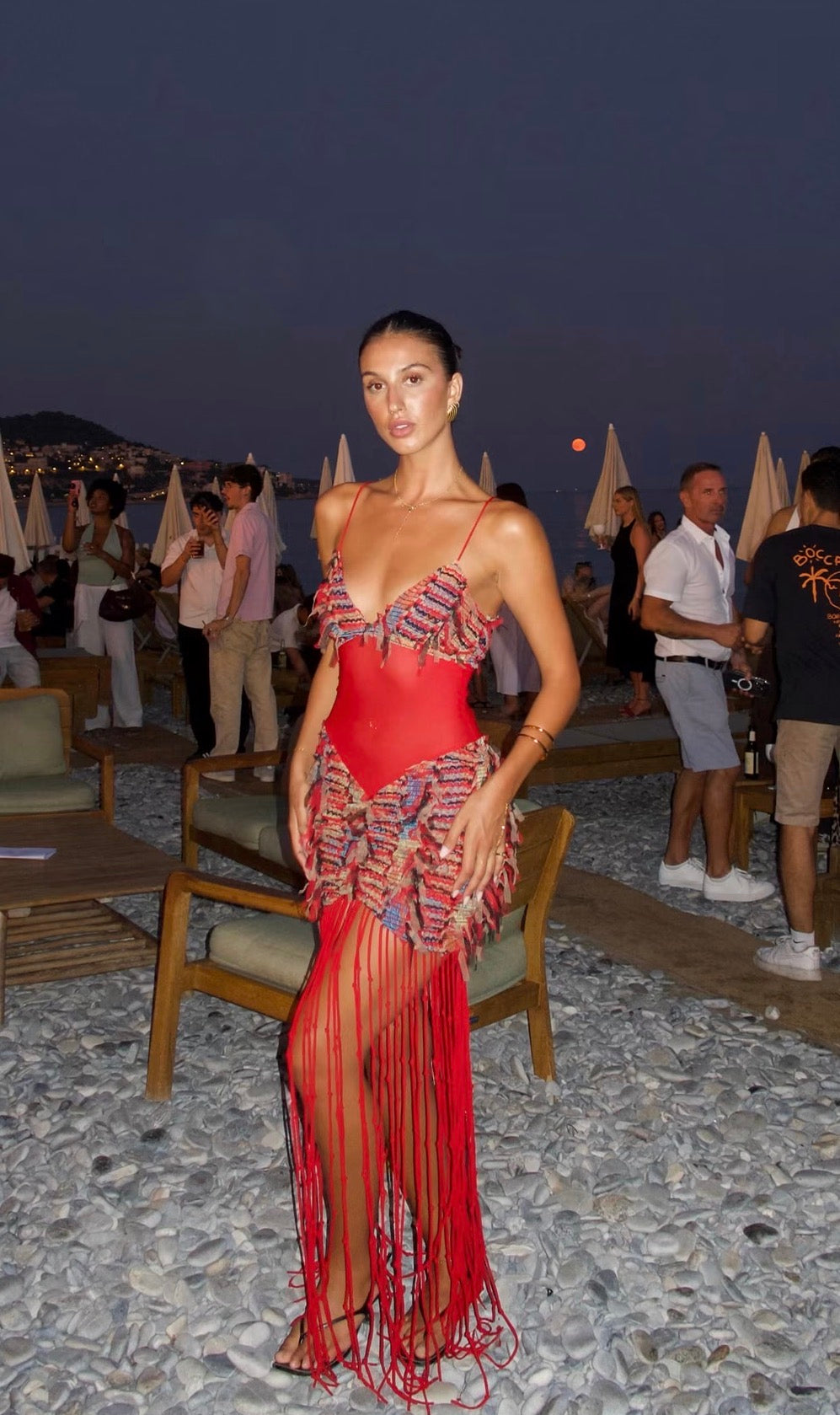 Woman wearing the Jefre Sei Carmen Dress in red, featuring a plunging V-neckline, sheer mesh bodice, multicoloured textured detailing across the bust and hips, and dramatic floor-length red fringe, photographed at a beachside venue at dusk.