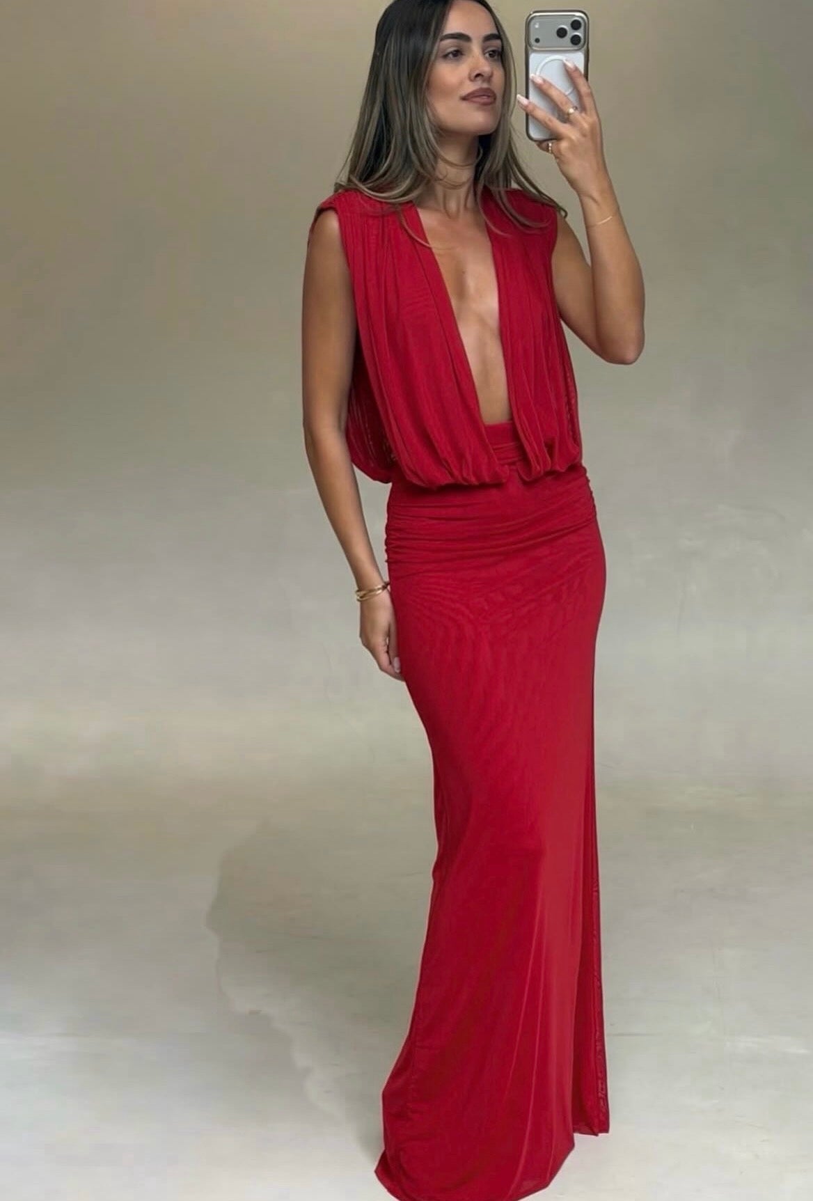 Woman wearing a vibrant red floor-length maxi dress featuring a deep plunging V-neckline, softly draped sleeveless bodice, gathered waist and fitted skirt, taking a mirror selfie against a neutral background.