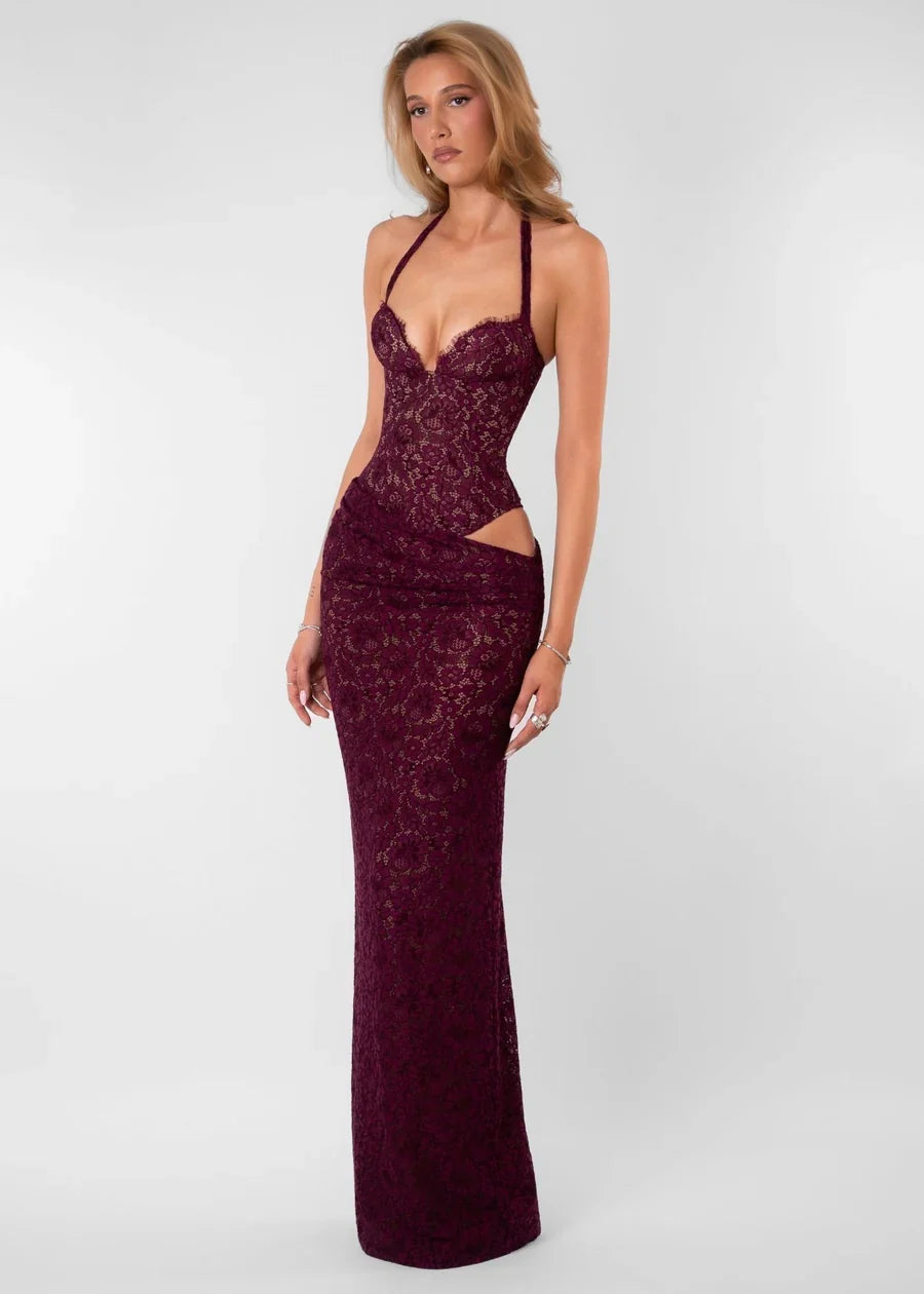 A woman modeling a floor-length, halter-neck gown in deep burgundy lace against a light gray studio background.