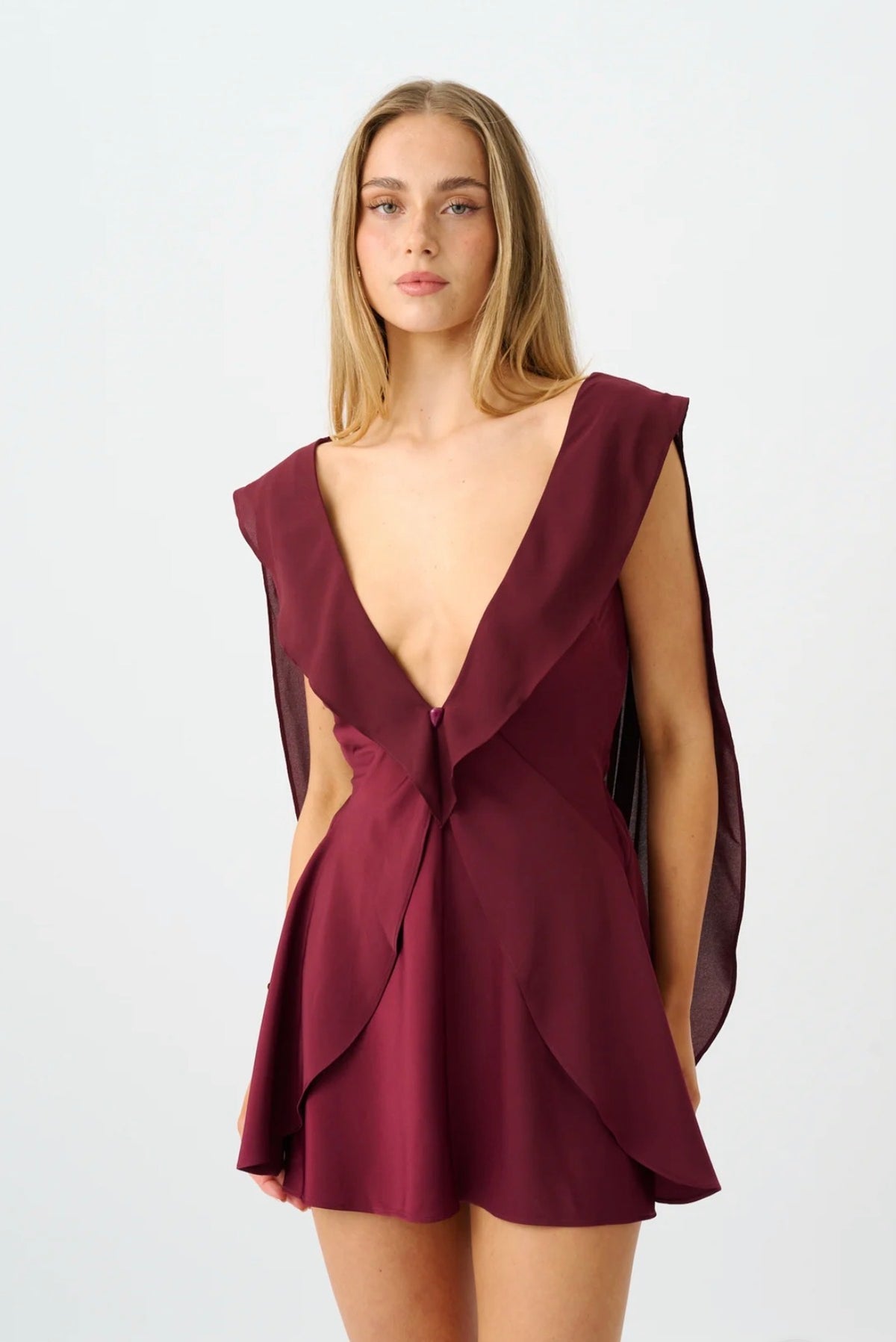 A woman modeling a sleeveless deep V-neck mini dress in burgundy with draped side panels and a wrap-style ruffle detail at the waist, photographed against a plain light gray studio background.
