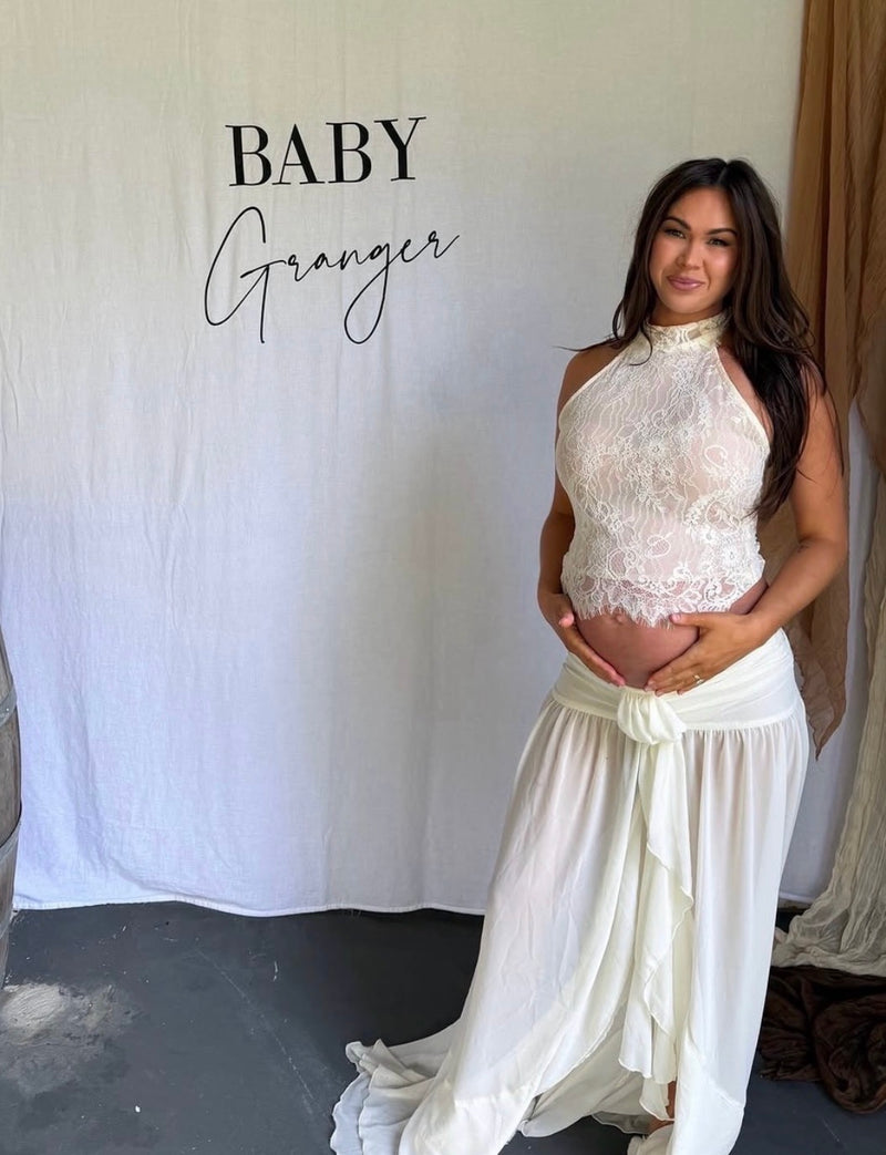 Pregnant woman in a white lace outfit standing against a white backdrop with 'BABY Granger' text. - @kristiegranger_
