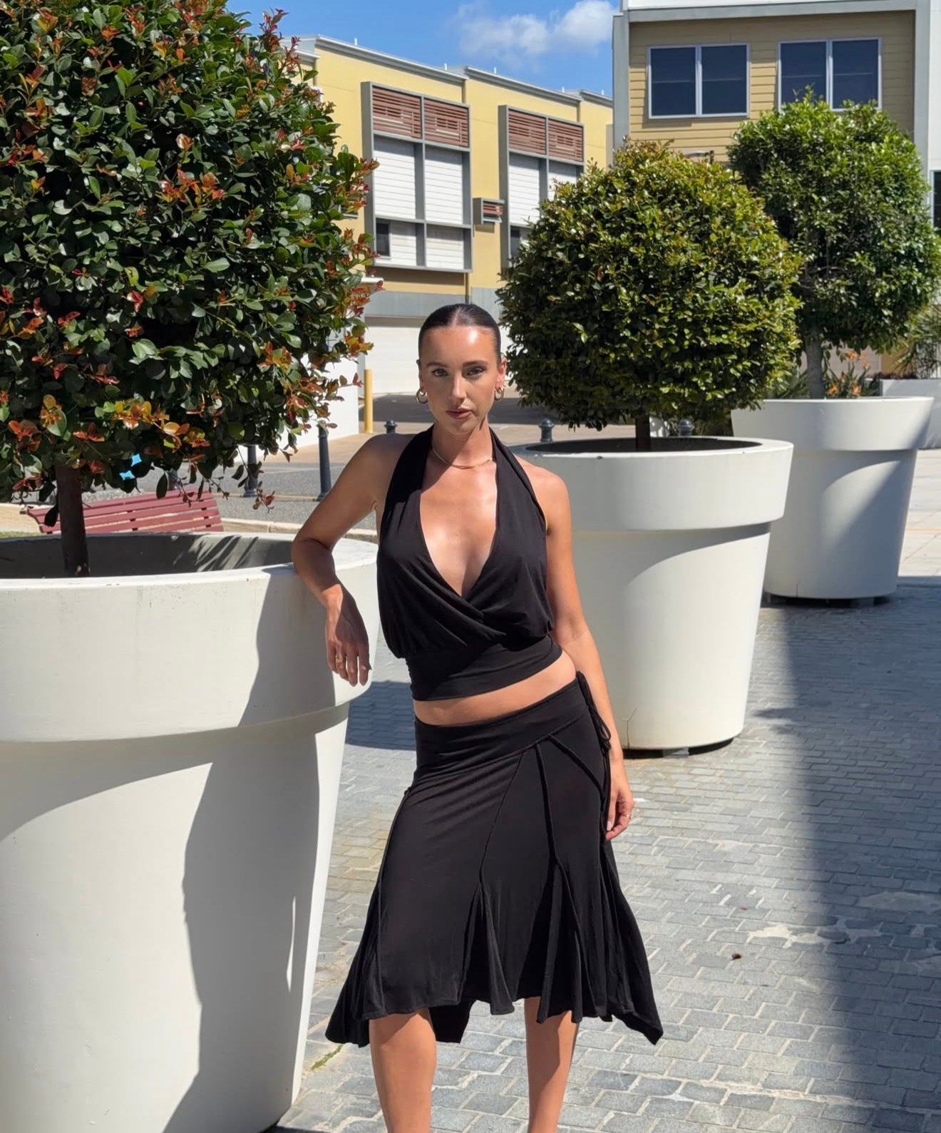 Woman in a black outfit standing outdoors with greenery and buildings in the background