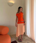 Woman in an orange top and beige skirt standing in a room with orange furniture and white curtains.