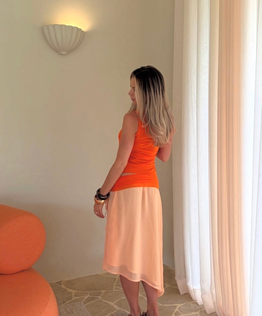 Woman wearing an orange top and peach skirt standing in a room with beige walls and a white curtain.