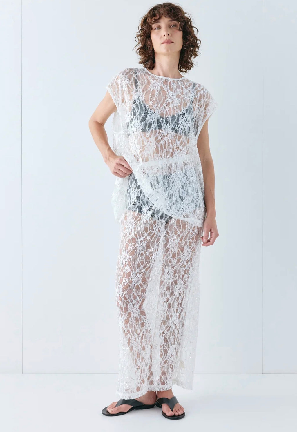 Model wearing the VRG GRL Devyn Sequin Set, featuring a sheer white lace top with delicate sequin embroidery and a matching midi skirt, styled over black undergarments with black sandals against a white studio background.