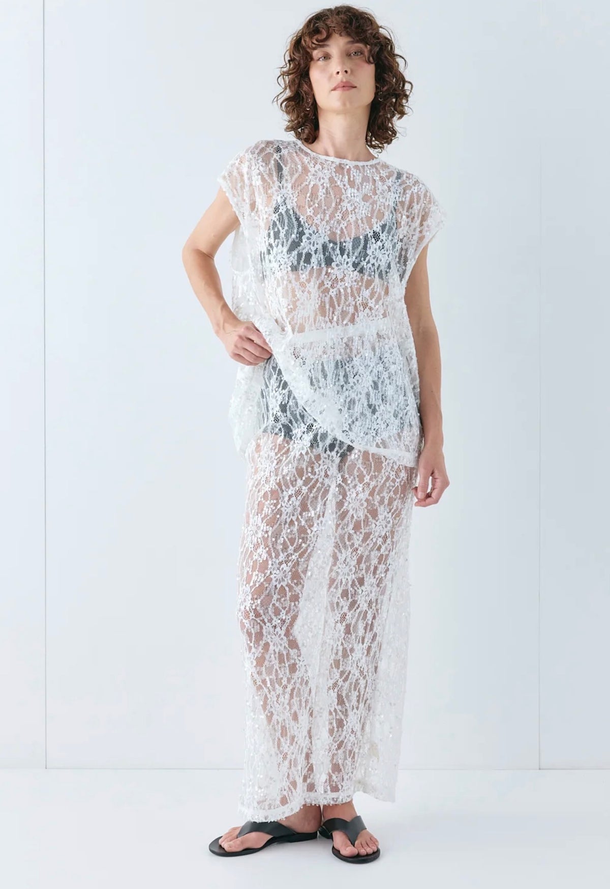 Model wearing the VRG GRL Devyn Sequin Set, featuring a sheer white lace top with delicate sequin embroidery and a matching midi skirt, styled over black undergarments with black sandals against a white studio background.
