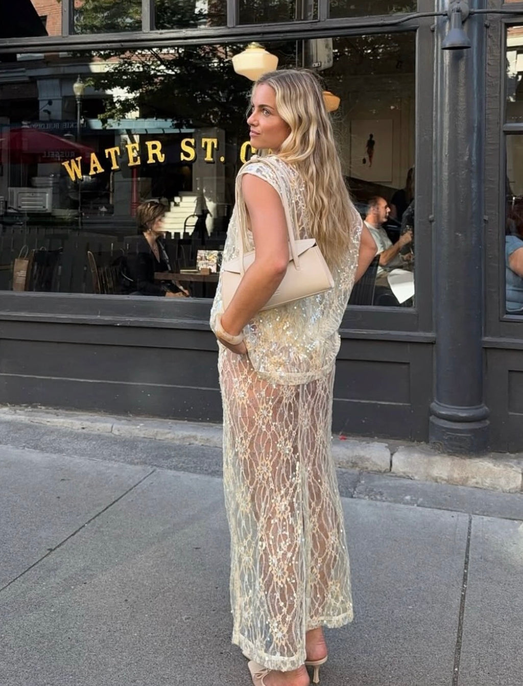 Woman modelling the VRG GRL Devyn Sequin Set, featuring a sheer sequin-embroidered top and matching lace midi skirt, styled with a cream shoulder bag and heels on a city street.