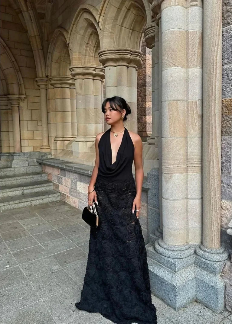 Woman wearing the black Arcina Ori Adriana Maxi Dress, featuring a plunging draped cowl neckline and textured rose appliqué A-line skirt, standing against sandstone arches.
