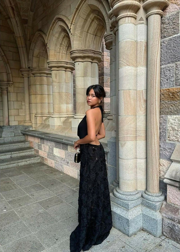 Woman modelling the black Arcina Ori Adriana Maxi Dress, showcasing its elegant open back, halter neckline and textured rose appliqué floor-length skirt, standing beside sandstone arches.