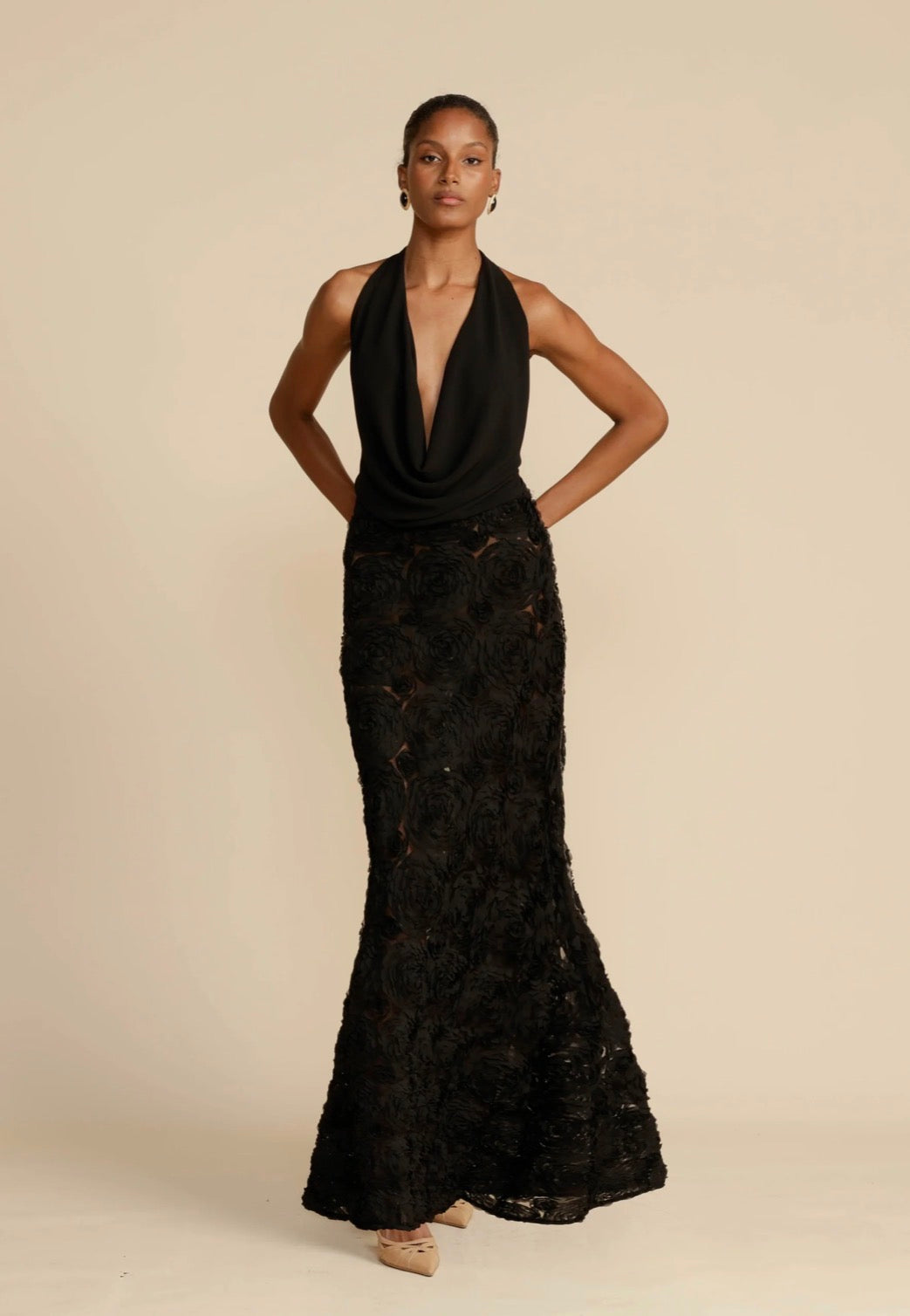 Model wearing the black Arcina Ori Adriana Maxi Dress, featuring a plunging cowl halter neckline, fitted drop waist and sheer rose appliqué A-line skirt, photographed against a neutral beige background.