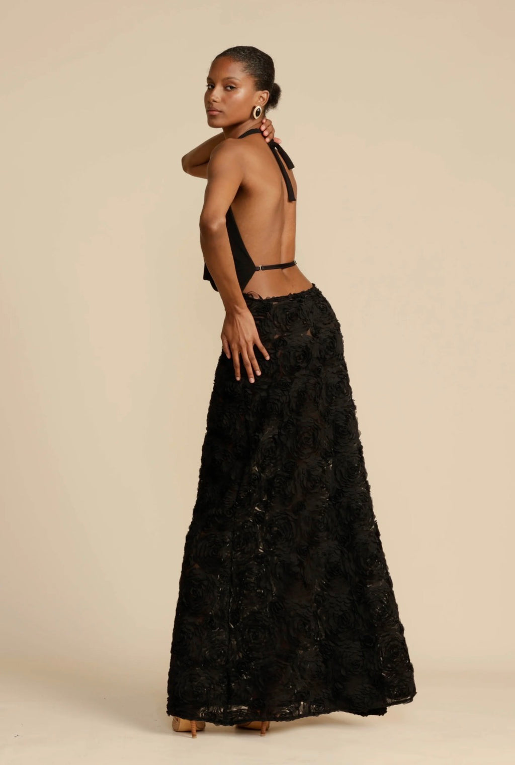 Model showcasing the back of the black Arcina Ori Adriana Maxi Dress, featuring an open-back halter design, adjustable waist strap and flowing rose appliqué A-line skirt against a neutral beige background.