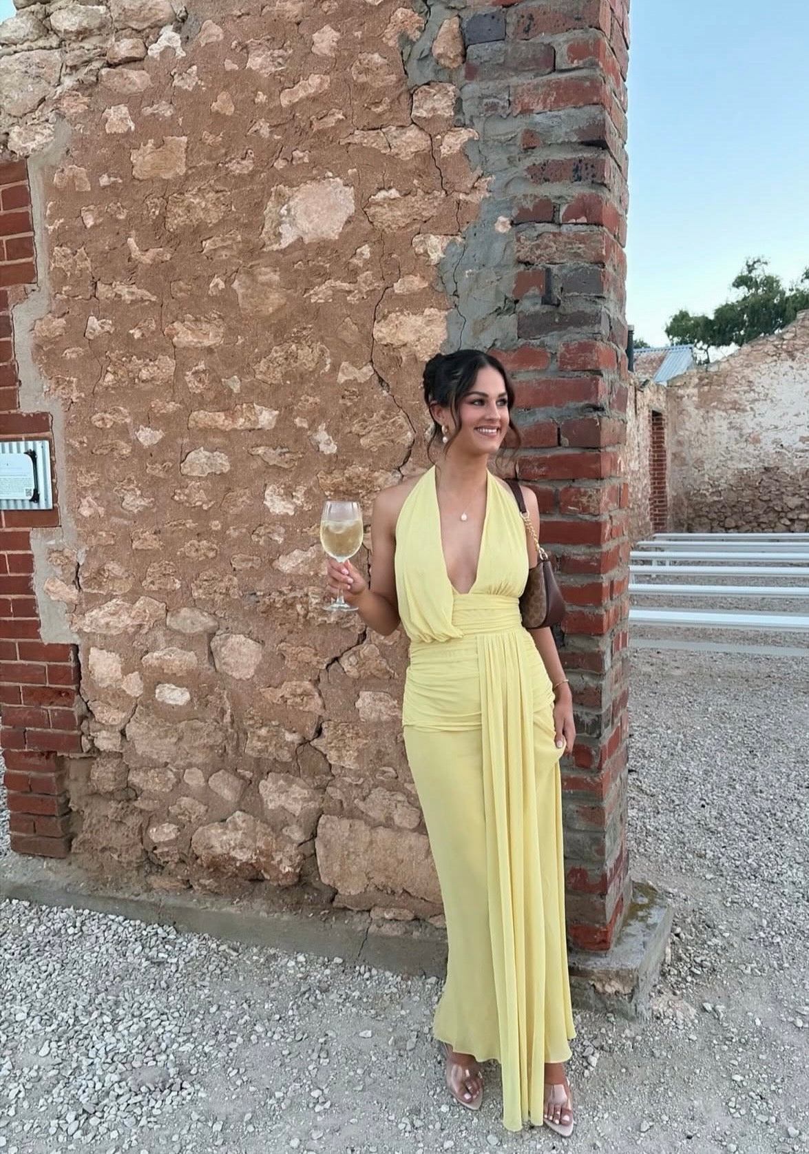 Woman in a yellow dress holding a glass of wine against a stone wall.