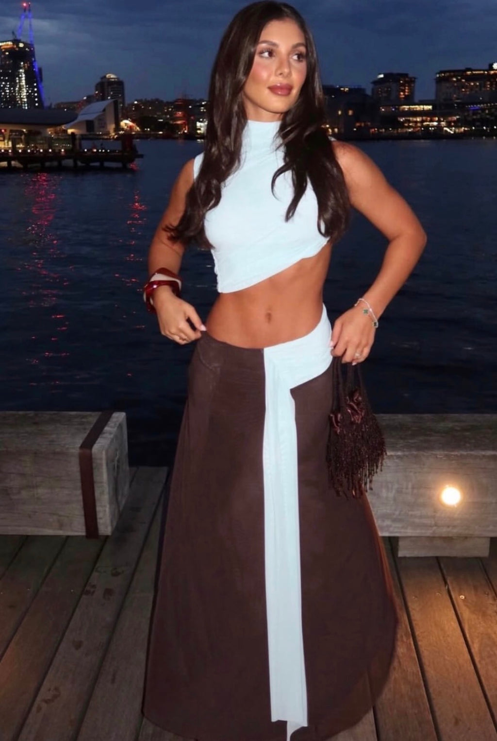 Woman in a white top and brown skirt standing on a dock with city skyline in the background