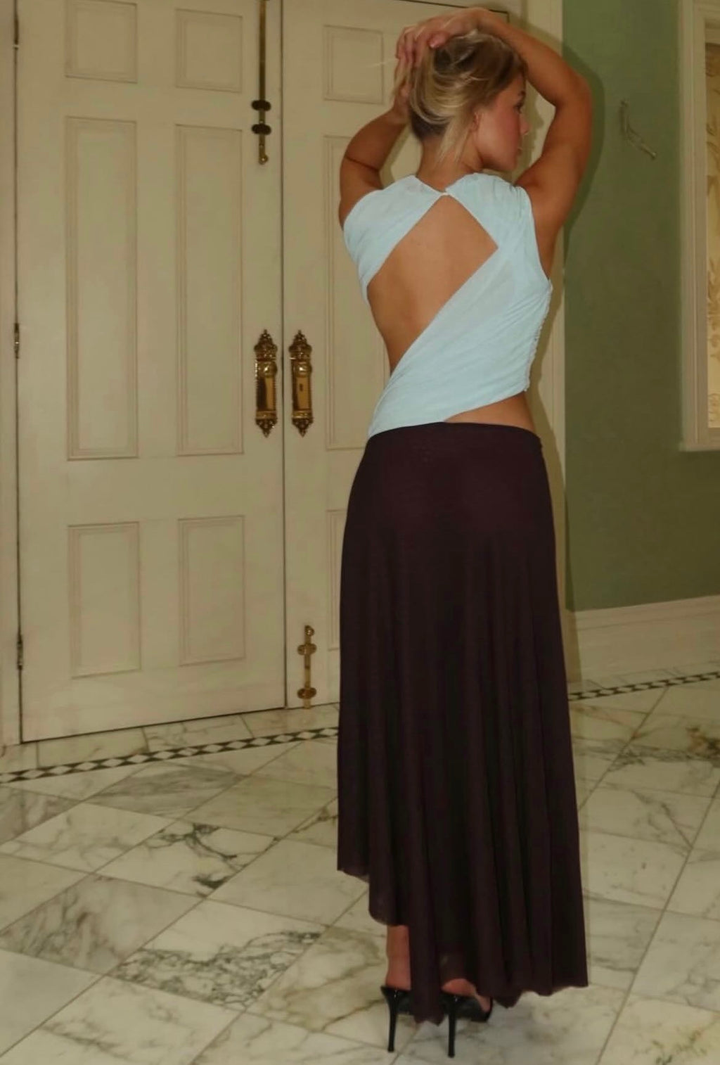 Woman in a light blue top and dark brown skirt standing in a room with marble flooring.