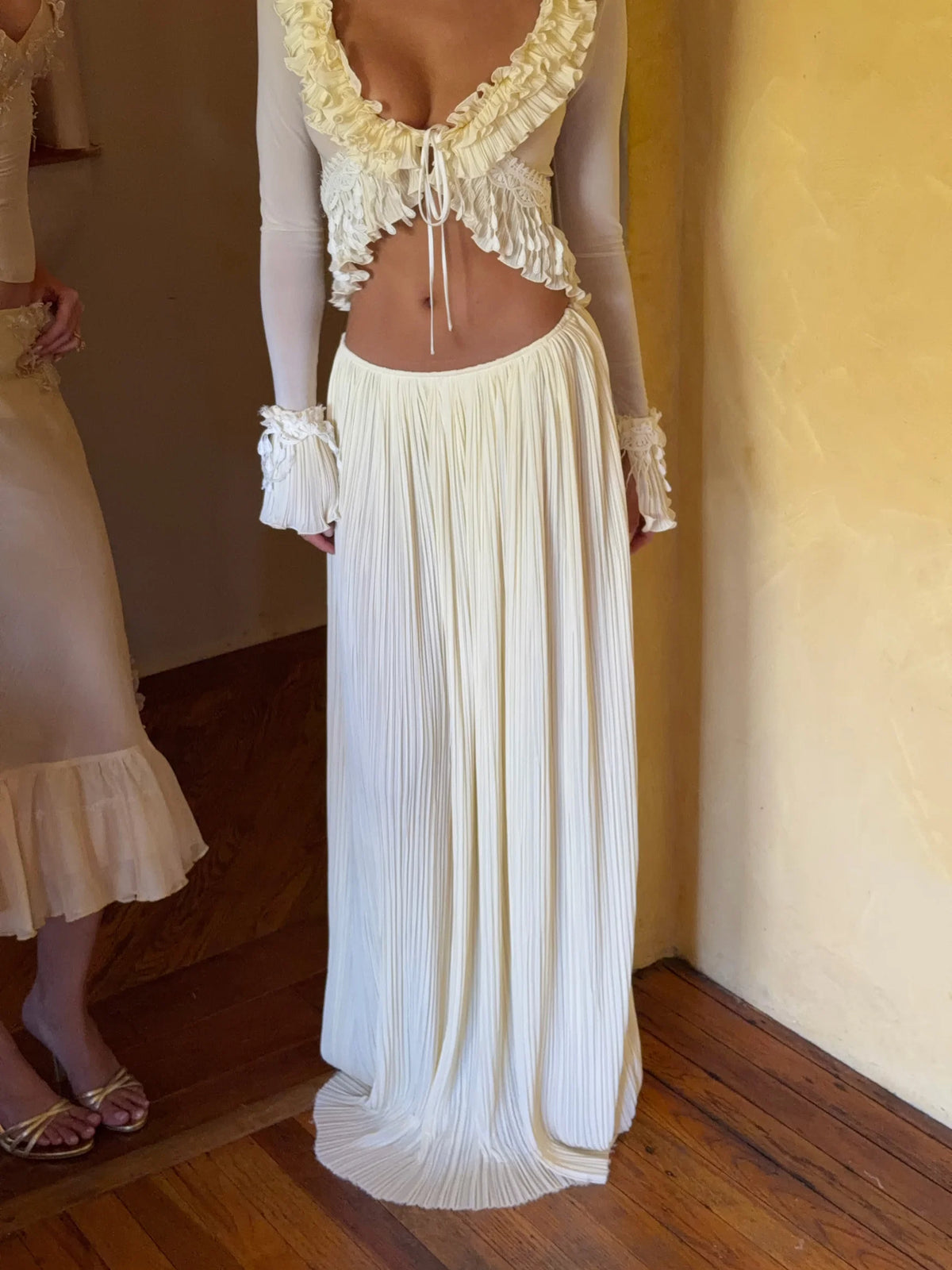 Front view of the Mali Marea Bellissima Set in ivory, featuring a plunging ruffled neckline, sheer long sleeves, delicate lace detailing, adjustable front tie and a flowing low-waisted pleated maxi skirt.