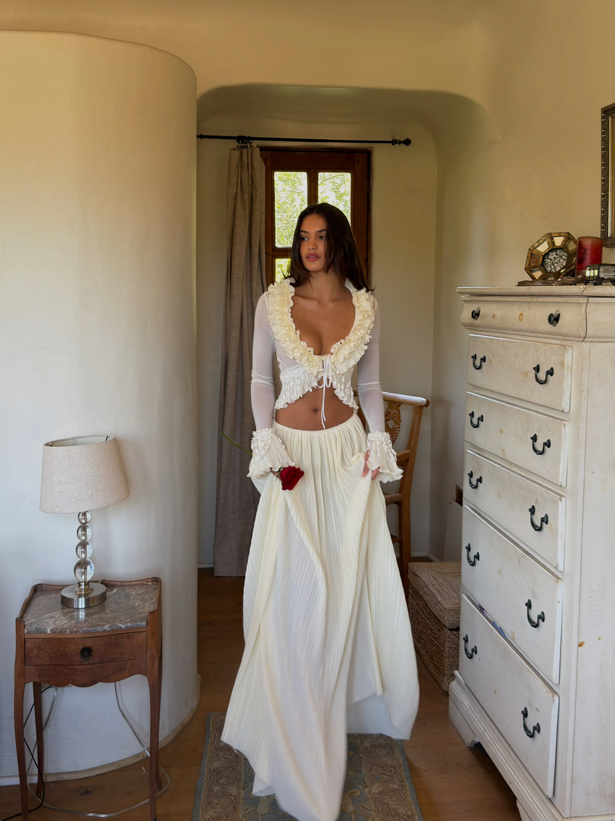 Model wearing the Mali Marea Bellissima Set in ivory, featuring a sheer long-sleeve crop top with cascading chiffon ruffles, lace detailing and a plunging neckline, paired with a flowing low-waisted pleated maxi skirt.