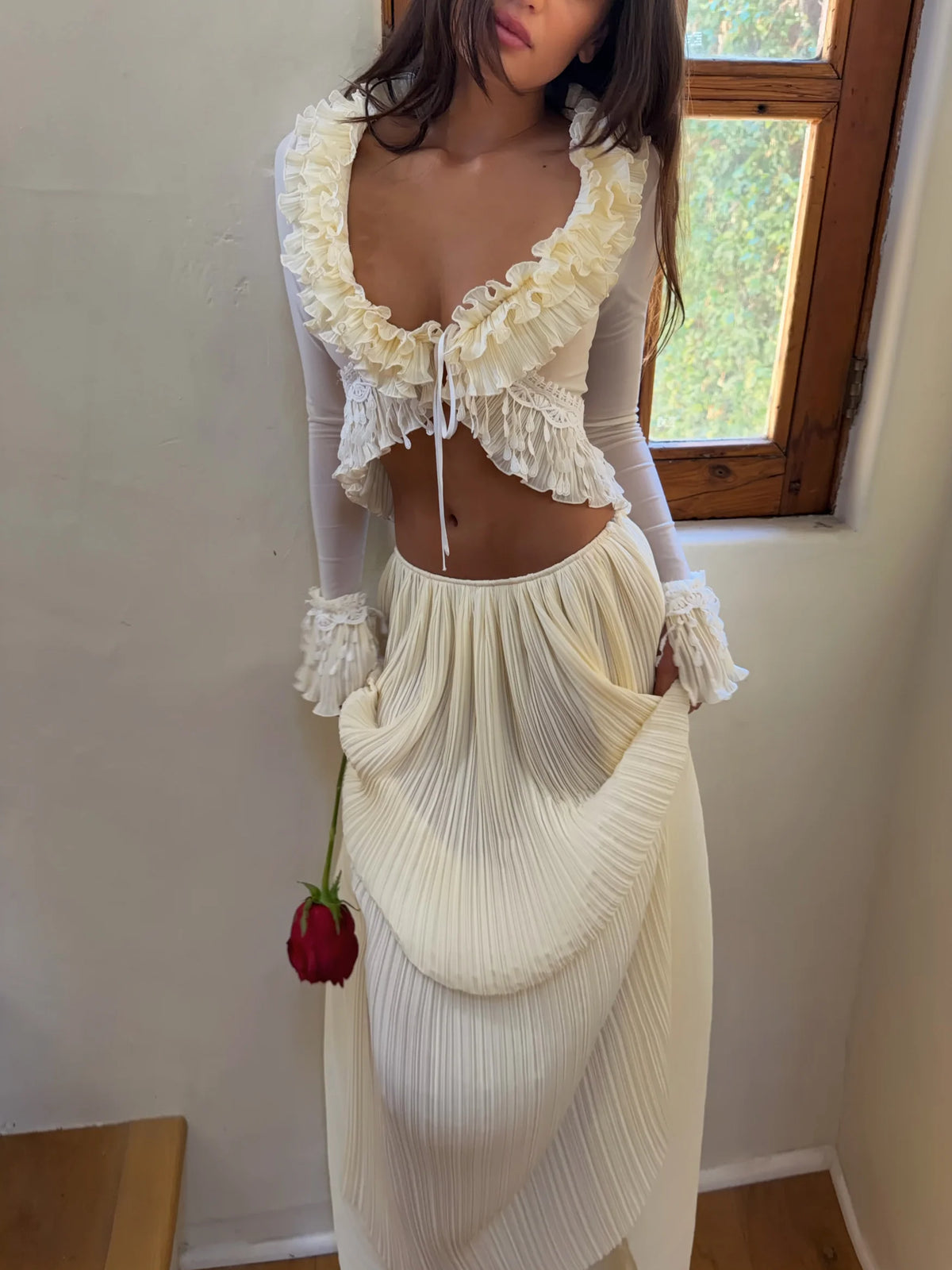 Model wearing the Mali Marea Bellissima Set in ivory, featuring a sheer long-sleeve crop top with cascading chiffon ruffles, lace detailing and a plunging neckline, paired with a flowing low-waisted pleated maxi skirt.