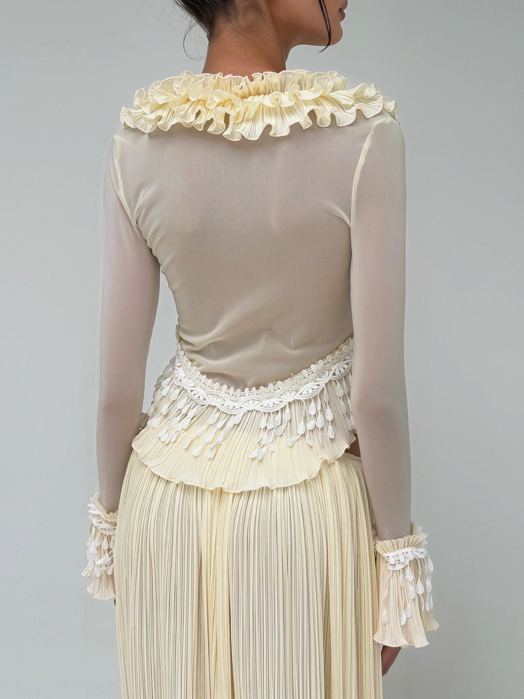 Back view of the Mali Marea Bellissima Set in ivory, featuring a sheer long-sleeve top with a pleated ruffle neckline, intricate lace detailing, cascading ruffled hem and matching pleated maxi skirt.