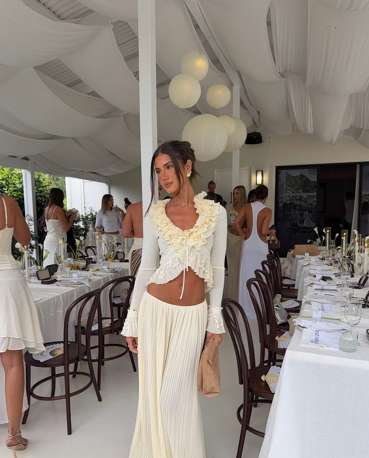 Model wearing the Mali Marea Bellissima Set in ivory, featuring a long-sleeve ruffled crop top with delicate lace detailing and a flowing pleated low-waisted maxi skirt, styled with a beige clutch at an elegant event venue.