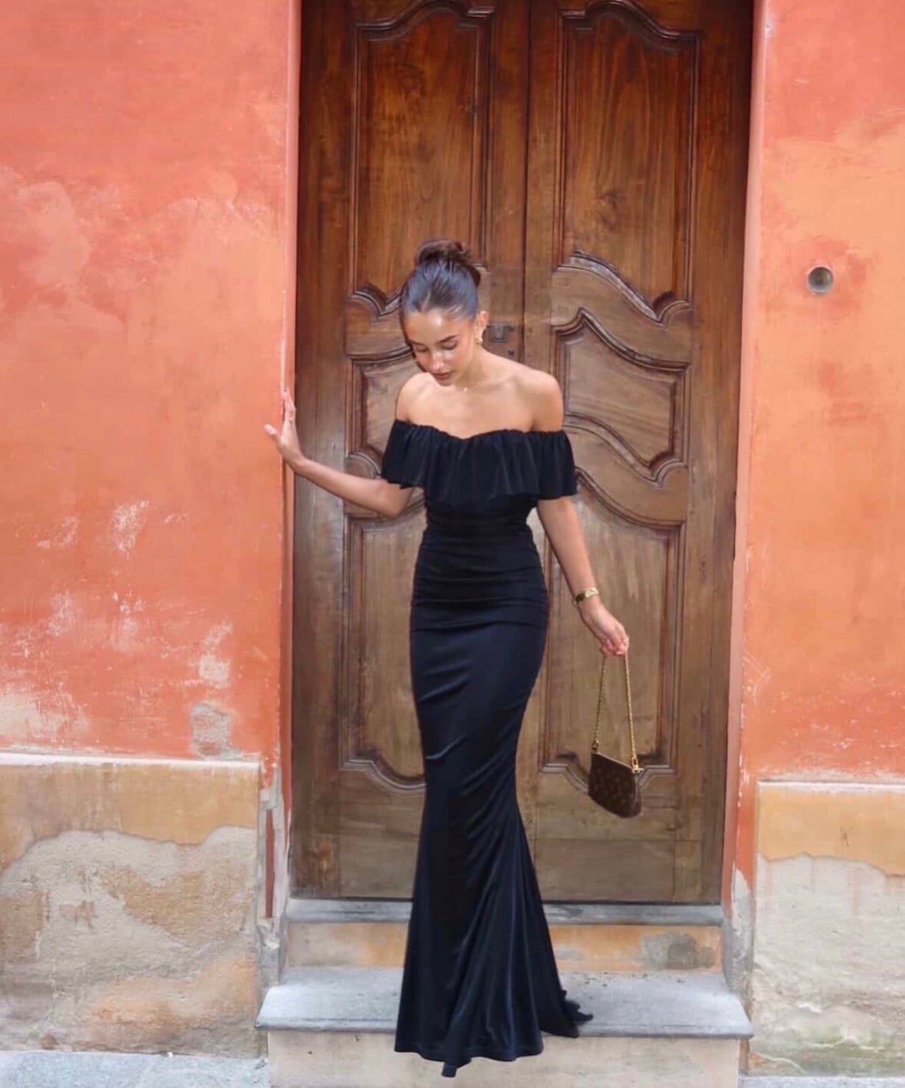 Woman in a black off-shoulder dress standing in front of a wooden door on a colorful wall.