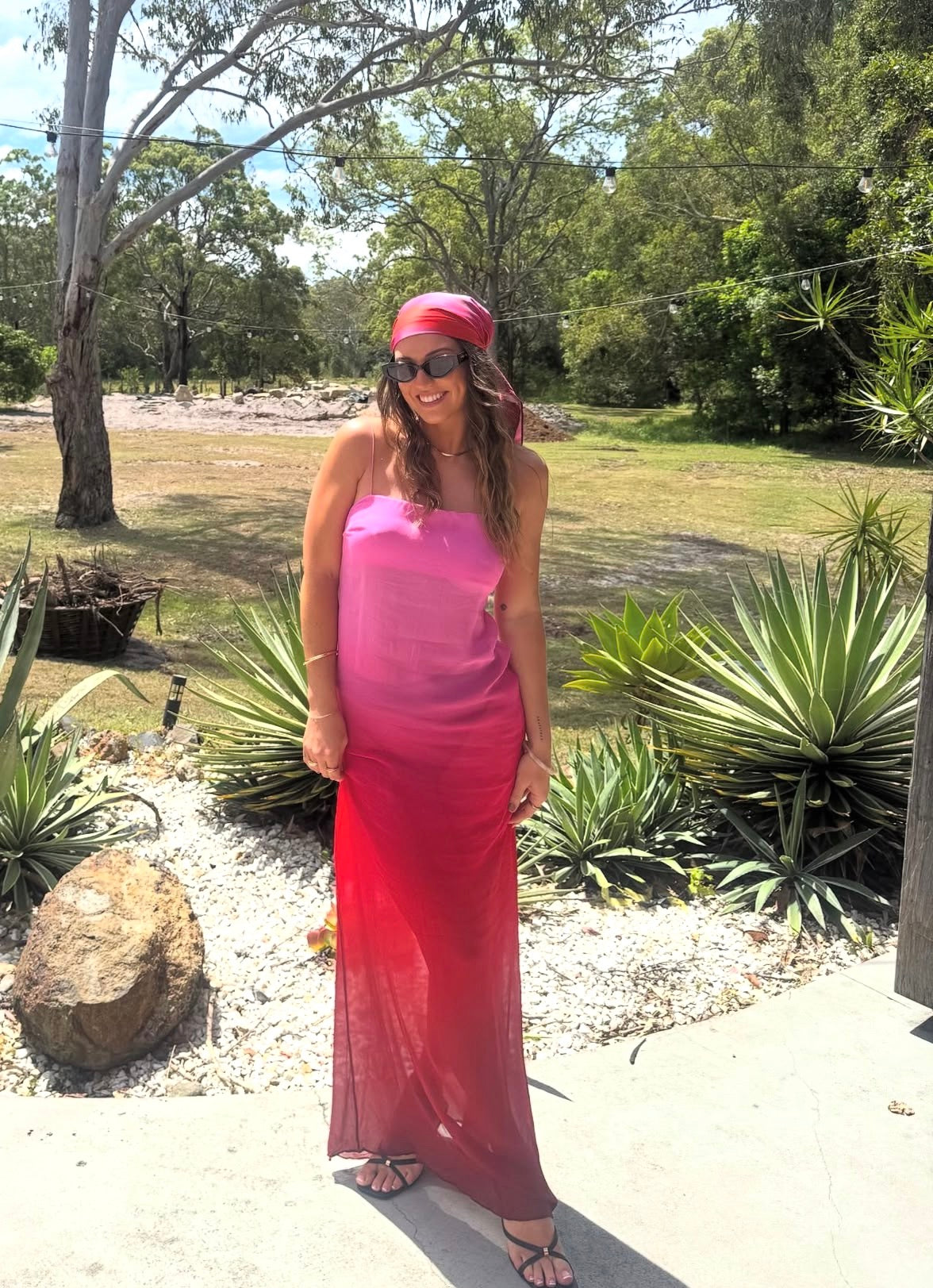 Woman in a pink dress and red headscarf standing outdoors with greenery in the background. - @_rachbell