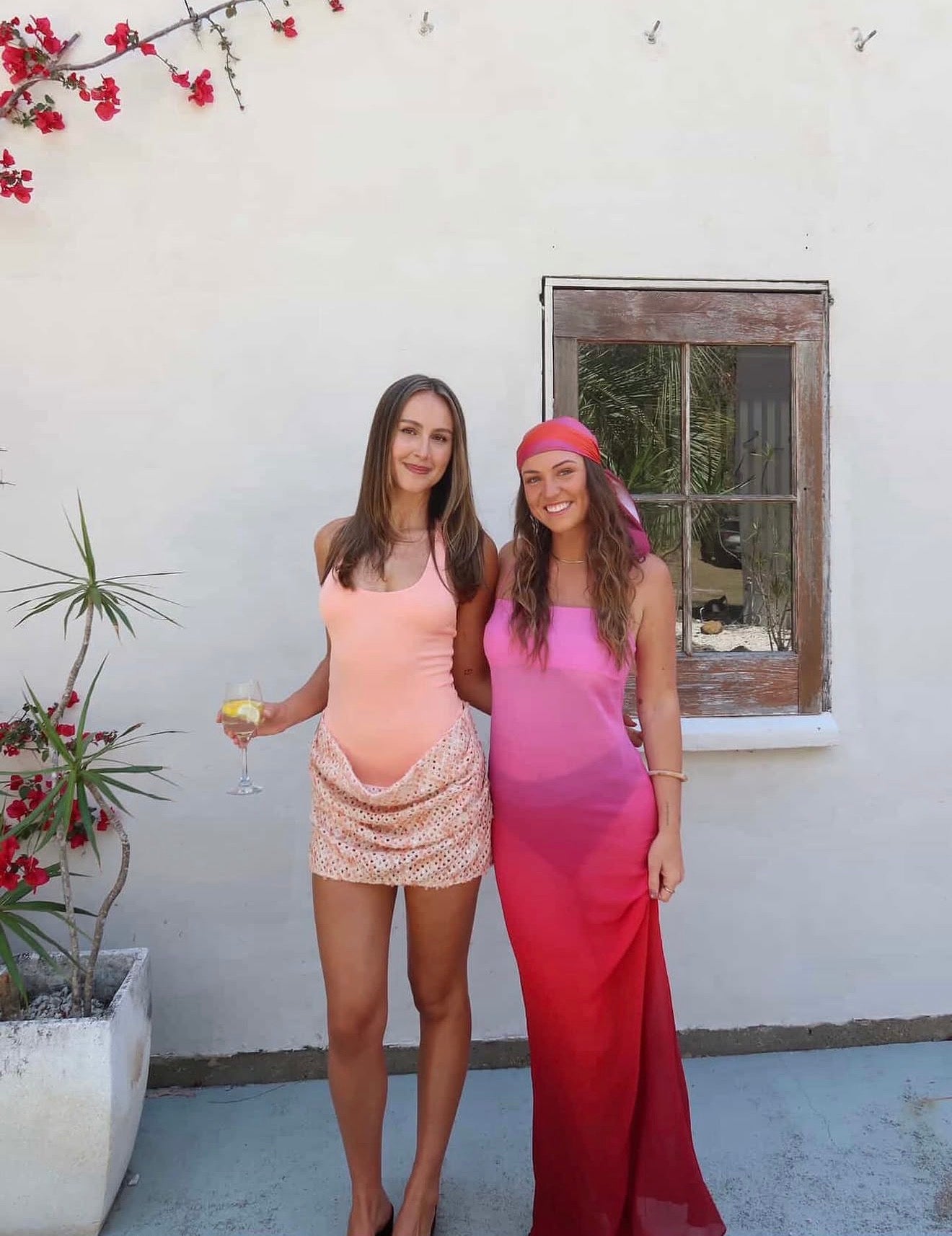 Two women standing outdoors, one in a pink dress and the other in a red dress, with a white wall and window in the background.