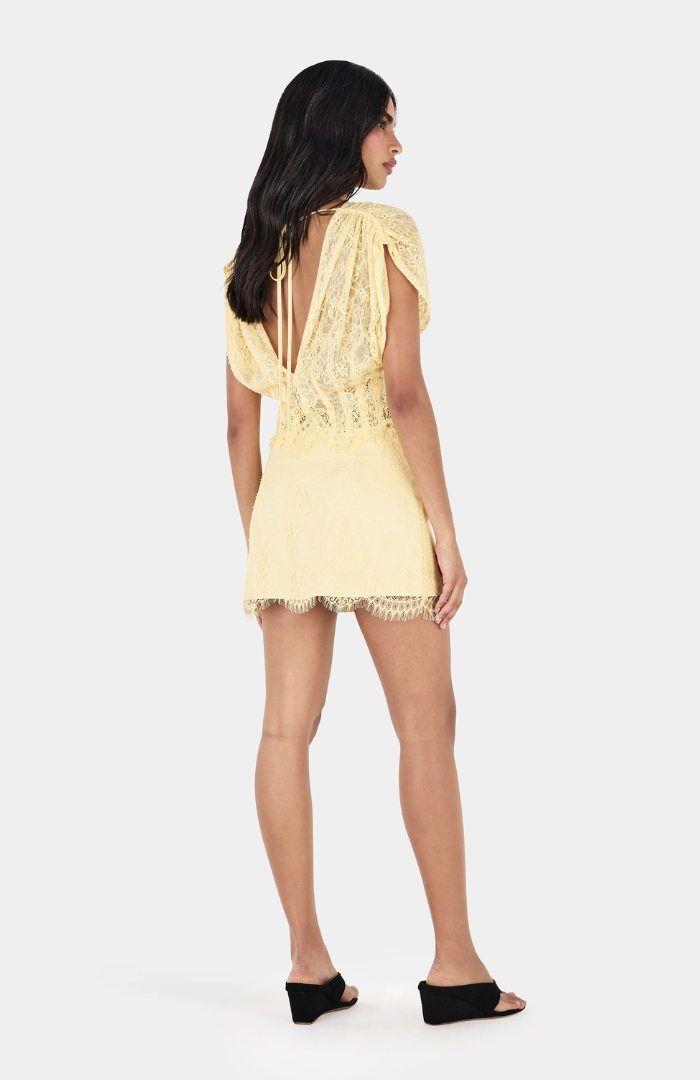Back view of a model wearing the Yasmin Lace Skirt and Ruby Lace Top matching set in butter yellow, featuring delicate floral lace, a deep V-shaped open back with adjustable ties, draped short sleeves and a fitted mini skirt with scalloped lace hemline, styled with black wedge heels against a white studio background.