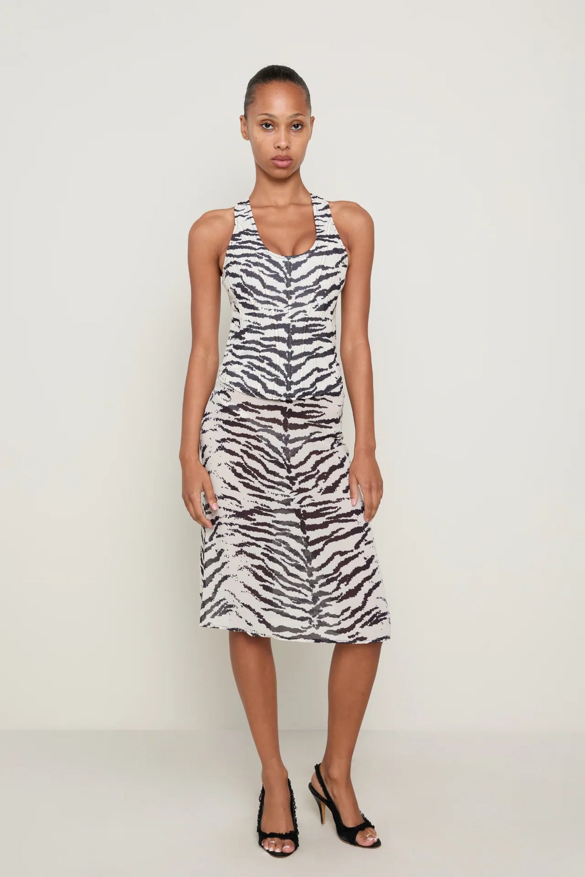 Front view of a model wearing the Miaou Annika Corset and Aimeé Skirt matching set in black and white zebra print, featuring a structured scoop-neck corset with wide straps, a fitted waist and a coordinating sheer midi skirt with a subtle flared hem, styled with black heels against a neutral studio background.