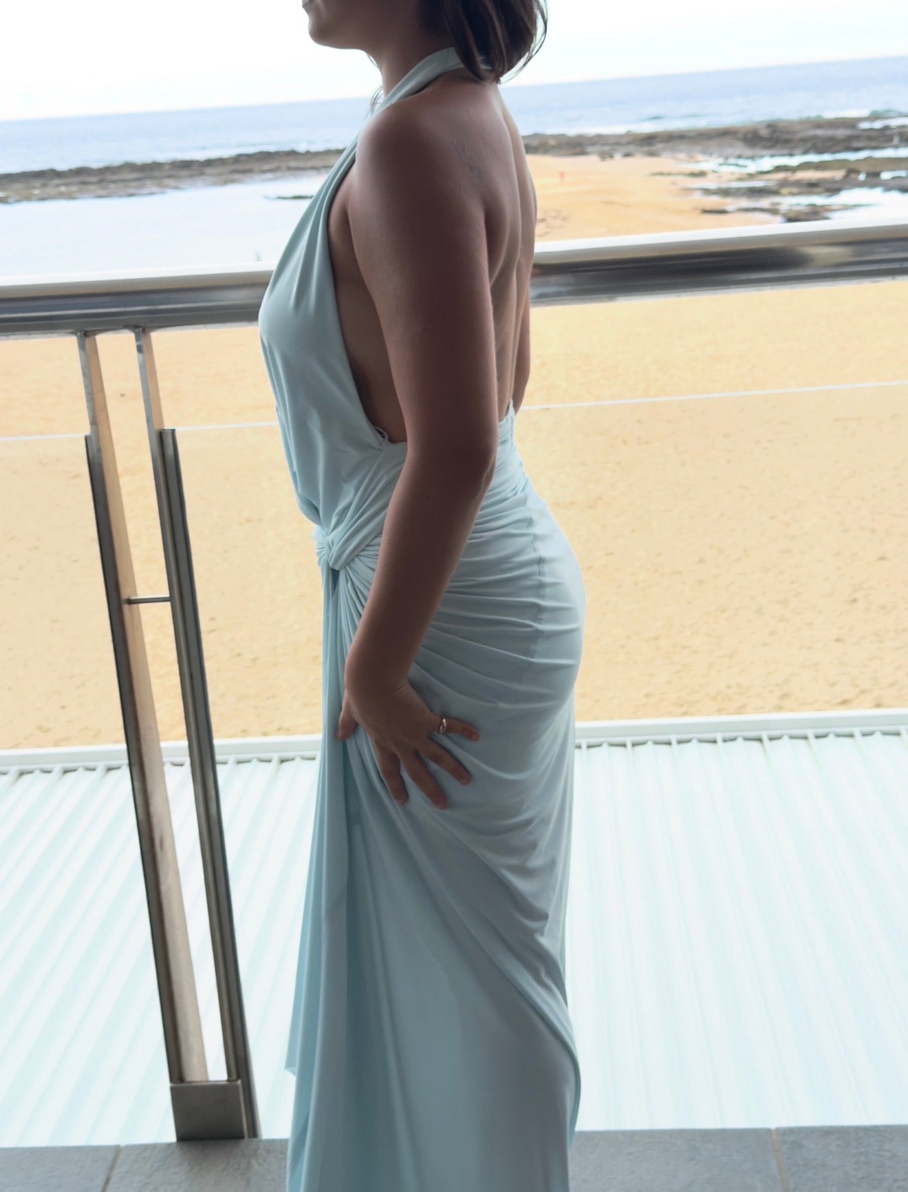 Woman in a light blue evening gown standing on a balcony with a beach view.