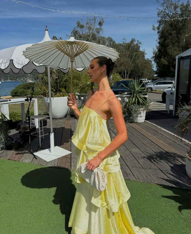 Woman in a yellow dress holding a drink on a deck with outdoor furniture and plants. - @alesha.beggs
