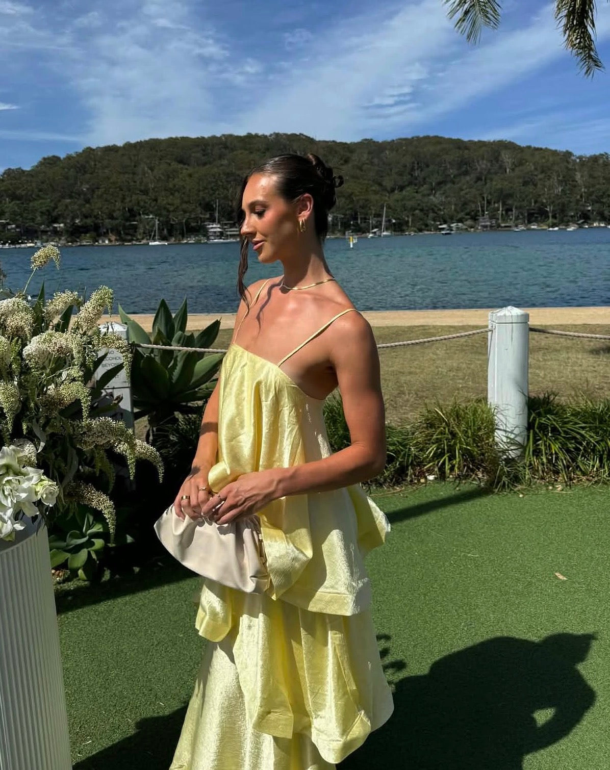 Woman in a yellow dress standing outdoors by a waterfront with greenery and water in the background.