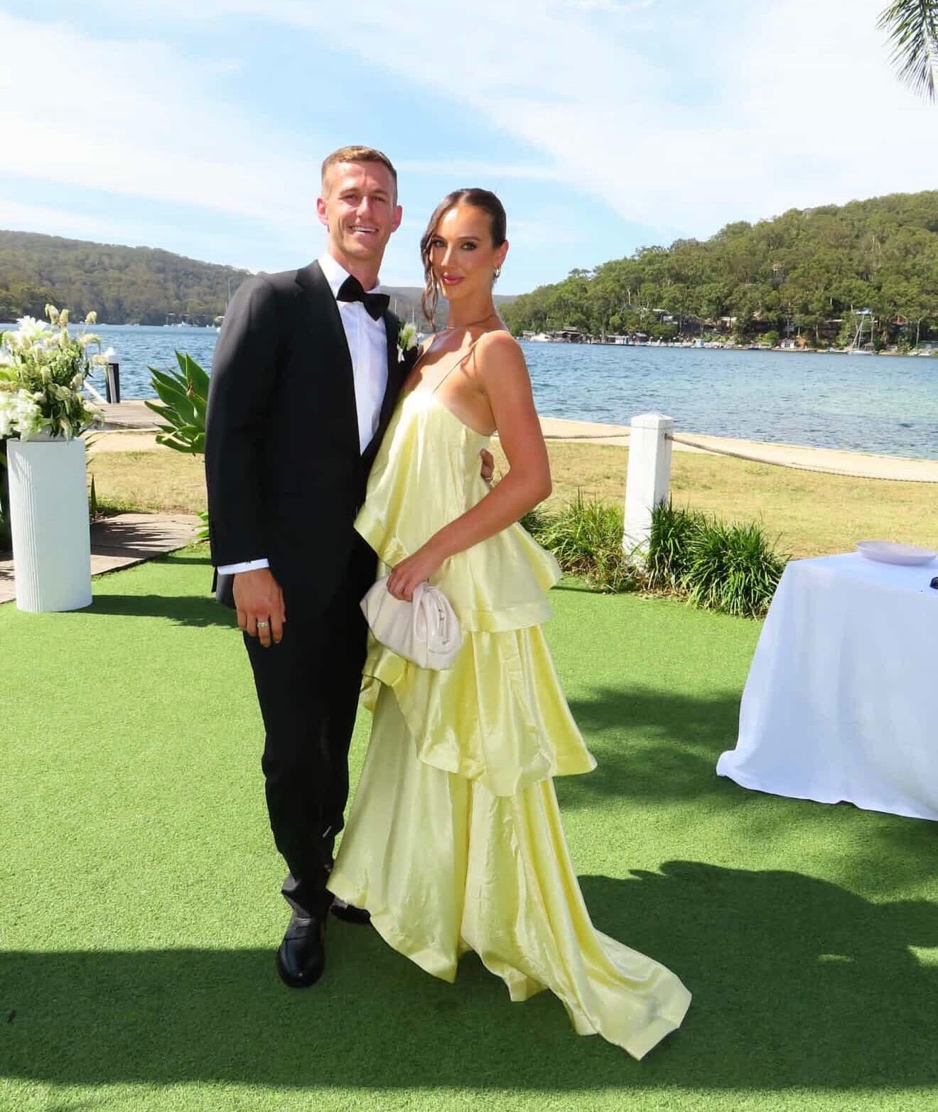 Man in a suit and woman in a yellow dress standing on grass with a lake and trees in the background